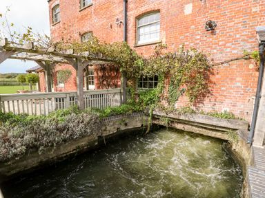 A brick building with a wooden pergola and climbing plants next to a water channel at The Happy Hut in Sopley near Christchurch