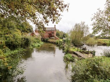 A river with green trees and bushes along the banks and a red brick building near the water at The Happy Hut in Sopley near Christchurch