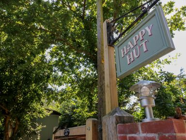 A wooden post with a hanging sign that reads happy hut near a metal outdoor light and green trees at The Happy Hut in Sopley near Christchurch