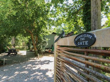 A wooden gate with a sign saying please close the gate overlooking a gravel path and a deck area with picnic table and trees at The Happy Hut in Sopley near Christchurch