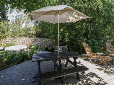 A wooden picnic table with an umbrella on a deck near two wooden lounge chairs and greenery at The Happy Hut in Sopley near Christchurch
