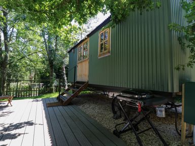 A green metal cabin with wooden door and windows on a gravel area next to a wooden deck with a barbecue grill nearby at The Happy Hut in Sopley near Christchurch