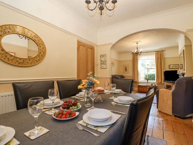 A dining table set with plates glasses and fruit in a room connected to a living area with chairs and a window at Green Gate Cottage in Reydon near Southwold