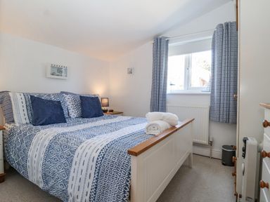 A bedroom with a bed with blue and white patterned bedding and cushions two bedside tables with lamps a window with patterned curtains a radiator and a chest of drawers at Green Gate Cottage in Reydon near Southwold