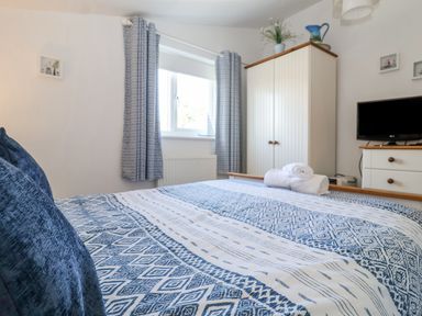 A bedroom with a bed with blue and white patterned bedding a window with blue curtains a white wardrobe a dresser with a TV and rolled towels on the bed at Green Gate Cottage Reydon near Southwold
