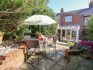 An outdoor patio with a table and chairs under an umbrella surrounded by plants and a brick house at Green Gate Cottage in Reydon near Southwold