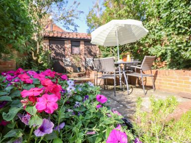 An outdoor patio with a table and four chairs under a large umbrella surrounded by flowering plants and greenery at Green Gate Cottage in Reydon near Southwold