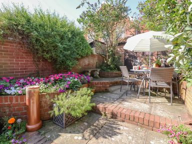 A garden patio with a table and chairs under an umbrella surrounded by brick walls and flower beds at Green Gate Cottage in Reydon near Southwold