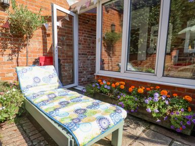 A sun lounger with patterned cushion on a brick patio surrounded by potted plants and flowers at Green Gate Cottage in Reydon near Southwold
