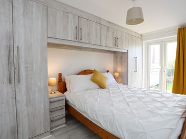 A bedroom with a wooden bed white bedding yellow pillow built in wardrobes bedside tables lamps and glass doors with yellow curtains at Seaside Sanctuary in Trearddur Bay
