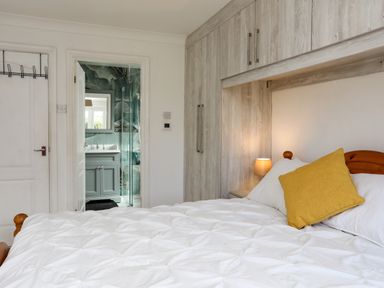 A bedroom with a bed yellow pillow bedside lamp wooden cabinets and a door leading to a bathroom at Seaside Sanctuary in Trearddur Bay