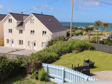 A yard with grass and overturned black chairs near a light blue wooden fence and a beige house with sea view at Seaside Sanctuary in Trearddur Bay