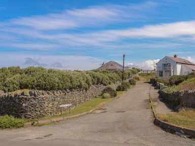 A road flanked by stone walls and bushes with houses in the background at Seaside Sanctuary in Trearddur Bay