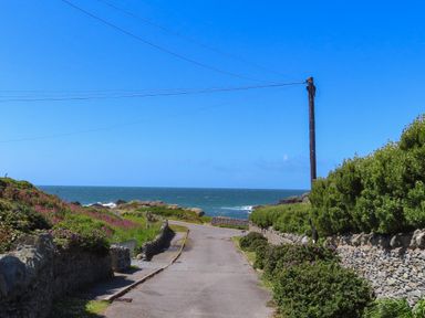 A coastal road with stone walls shrubs and flowers leading to the sea under a blue sky