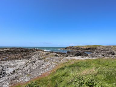 Rocky coastline with grassy foreground and ocean under blue sky at Seaside Sanctuary in Trearddur Bay
