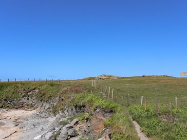 A rocky coastline with a grassy cliff and a wire fence under a clear blue sky at Seaside Sanctuary in Trearddur Bay