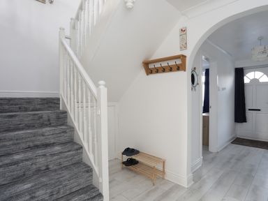 A hallway with stairs carpeted in grey and a wooden shoe rack under the stairs at Seaside Sanctuary in Trearddur Bay
