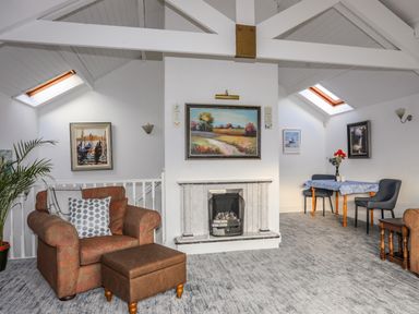 A living room with an armchair and ottoman a fireplace a dining table with two chairs and paintings on the wall at Seaside Sanctuary in Trearddur Bay