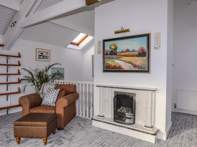 A sitting area with an armchair and ottoman next to a fireplace with a landscape painting above at Seaside Sanctuary in Trearddur Bay