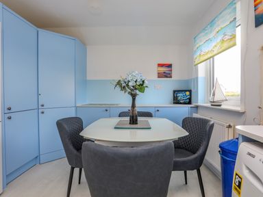 A dining area with a white table and gray chairs next to blue kitchen cabinets and a window with a painted blind at Seaside Sanctuary in Trearddur Bay