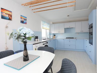 A kitchen with light blue cabinets a dining table with a vase of flowers and dark chairs at Seaside Sanctuary in Trearddur Bay