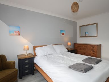 A bedroom with a bed white bedding two gray towels bedside tables lamps a wooden dresser mirror and a brown armchair at Seaside Sanctuary in Trearddur Bay