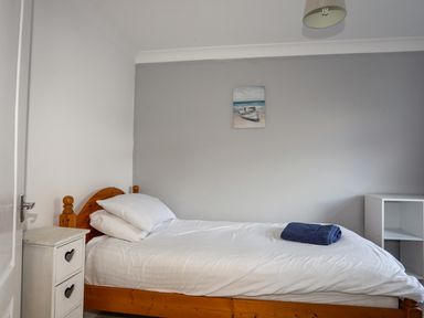 A bedroom with a single wooden bed with white bedding and two pillows a small white bedside table with heart-shaped cutouts and a small white shelf unit on the right wall painting of boats on the gray wall at Seaside Sanctuary in Trearddur Bay