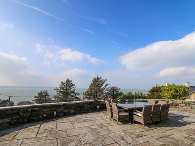 An outdoor patio with a table and chairs overlooking trees and the sea at Mynydd ar Mor in Barmouth
