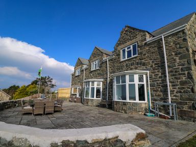 An outdoor patio area with stone paving and wicker dining furniture outside a stone house with large windows at Mynydd ar Mor in Barmouth