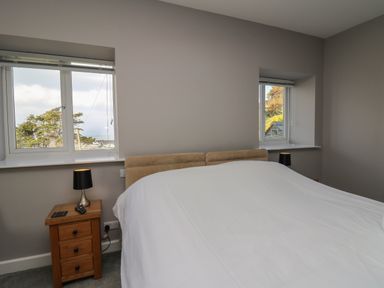A bedroom with a bed covered in a white sheet two wooden nightstands each with a lamp and two windows showing trees outside at Mynydd ar Mor in Barmouth