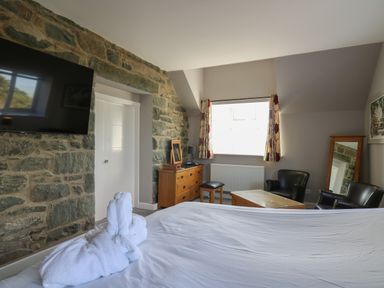 A bedroom with a stone wall, a mounted television, a wooden dresser, a window with curtains, two black armchairs, a wooden coffee table, and a bed with towels on it at Mynydd ar Mor in Barmouth