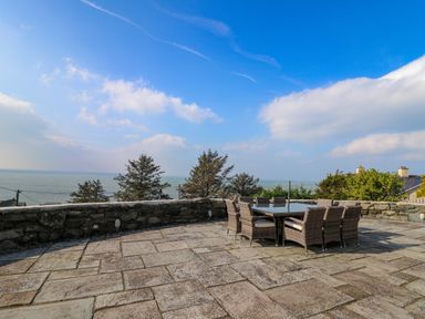 An outdoor stone patio with a glass table and wicker chairs overlooking the sea at Mynydd ar Mor in Barmouth