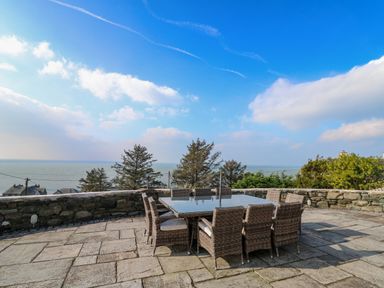 An outdoor patio with a stone floor a glass table and wicker chairs with a view of trees and the sea at Mynydd ar Mor in Barmouth