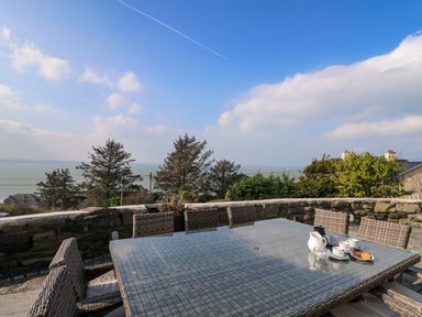 An outdoor patio with a glass table, wicker chairs, a tea set with cups and biscuits, overlooking trees and the sea at Mynydd ar Mor in Barmouth