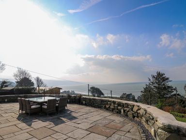 A stone patio with a glass table and wicker chairs overlooking the sea at Mynydd ar Mor in Barmouth