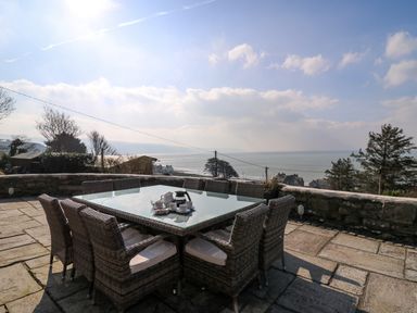 Outdoor patio with wicker chairs and a glass table overlooking the sea at Mynydd ar Mor in Barmouth