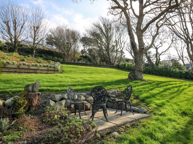A garden with a metal table and three chairs on a stone patio surrounded by grass and trees at Mynydd ar Mor in Barmouth