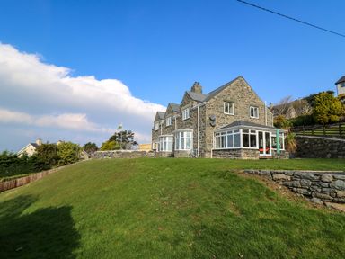 A stone house with multiple windows on a grassy hill under a blue sky at Mynydd ar Mor in Barmouth