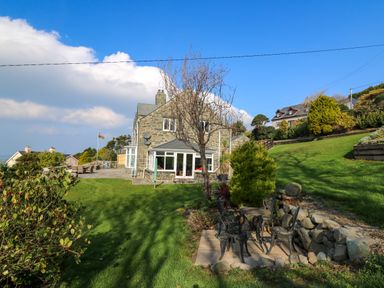 A stone house with a patio and garden furniture on a grass lawn with trees and bushes at Mynydd ar Mor in Barmouth