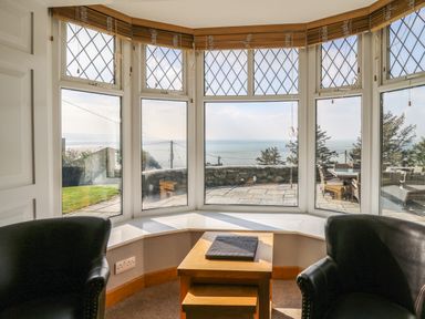 A sitting area with two black chairs and a wooden table by large bay windows overlooking a stone patio and sea at Mynydd ar Mor in Barmouth