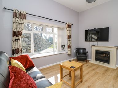 A living room with a black sofa with red and yellow cushions a wooden table a black armchair a TV above a fireplace and a window with patterned curtains at Mynydd ar Mor in Barmouth