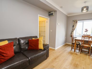 A living room with a black leather sofa with red and yellow cushions next to a wooden dining table with four chairs near a window at Mynydd ar Mor in Barmouth