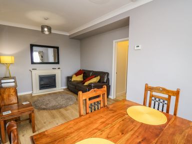 A living room with a wooden dining table and chairs leather sofa with cushions a fireplace a round rug and a lamp at Mynydd ar Mor in Barmouth
