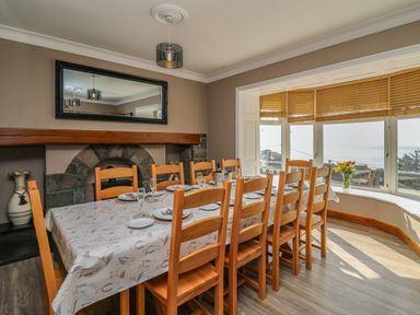 A dining room with a long table set for twelve people next to a bay window with wooden blinds and a view of the sea at Mynydd ar Mor in Barmouth