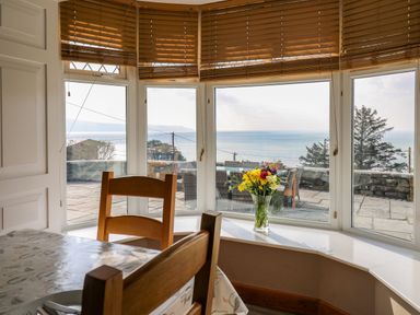 A dining area with wooden chairs and a table near a bay window with flowers on the windowsill overlooking the sea at Mynydd ar Mor in Barmouth