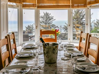 A dining table set with plates glasses and cutlery near large windows overlooking trees and the sea at Mynydd ar Mor in Barmouth