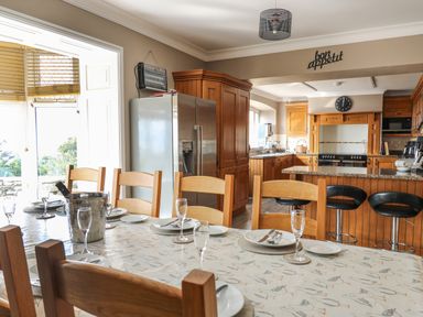 A dining table set with plates and glasses next to a kitchen with wooden cabinets and a refrigerator at Mynydd ar Mor in Barmouth