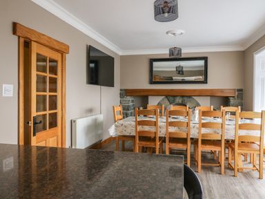 A dining room with a wooden table and chairs a wooden door a wall-mounted tv and a stone fireplace at Mynydd ar Mor in Barmouth