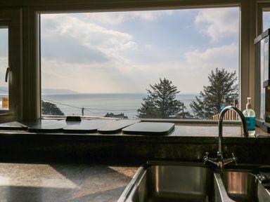A kitchen sink with a faucet and soap dispenser next to a window showing a sea view with trees and houses at Mynydd ar Mor in Barmouth