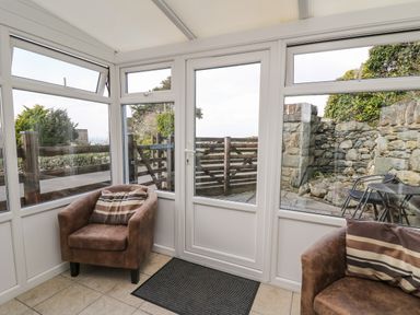 A conservatory with two brown armchairs and a striped cushion beside a glass door and windows showing a wooden gate and stone wall at Mynydd ar Mor in Barmouth